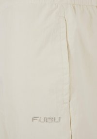 Șorturi de culoare bej deschis, confecționate dintr-un material moale, cu o textură subtilă, talie elastică și un logo brodat "FUBU" pe piciorul stâng.