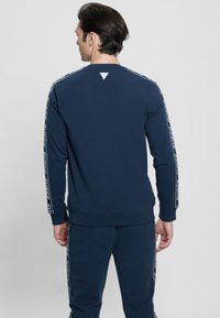 Felpa blu navy con collo a giro, polsini a coste e grafiche del logo "Guess" lungo le maniche. Tessuto morbido, design minimale.