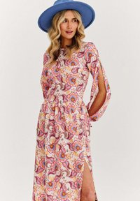 Květinové maxi šaty v světle růžové barvě s fialovými a oranžovými vzory, s dlouhými rukávy s výřezy a bočním rozparkem. Modrý klobouk s širokým krempou.