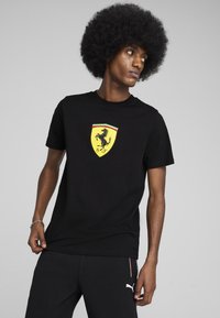 Čierne tričko s krátkymi rukávmi a bežným strihom, na ktorom je žlté logo Ferrari so skákajúcim koňom a zeleno-červeno-bielymi akcentmi na erbe.