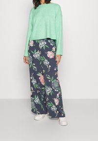 Kobieta w miętowozielonym swetrze, szerokich granatowych spodniach w duży kwiatowy wzór oraz białych butach slip-on, stojąca na jednolitym tle.