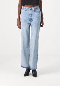 Jeans a gamba larga di un blu chiaro, tessuto denim con una finitura sbiadita e un sottile motivo perforato, abbinati a scarpe nere con tacco alto.