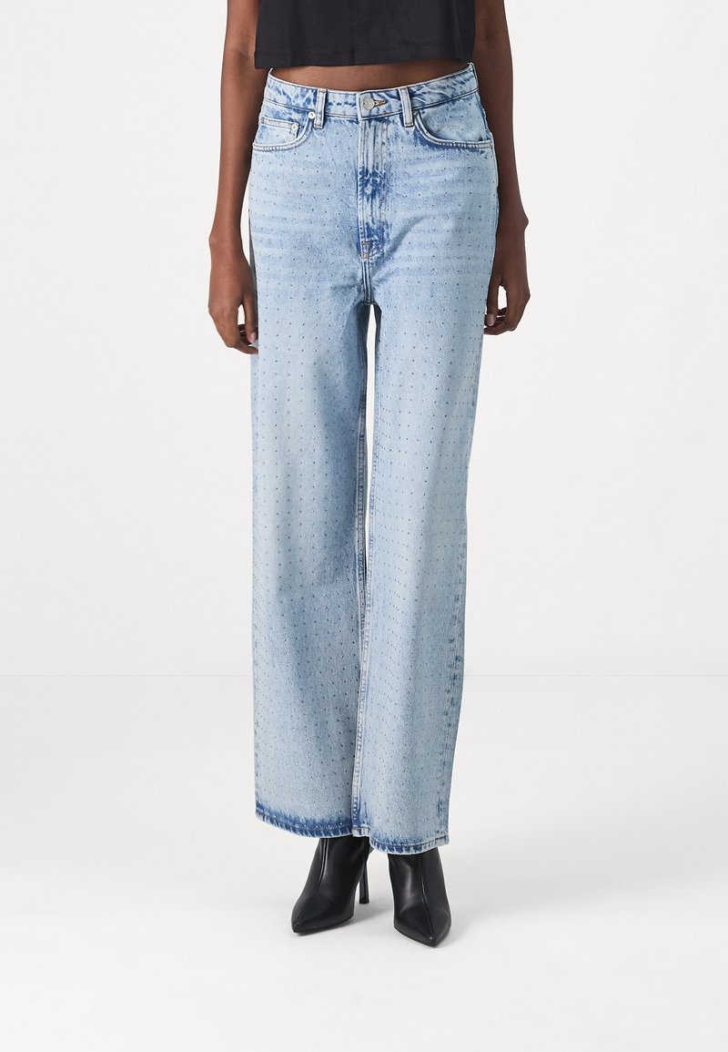 Jeans a gamba larga di un blu chiaro, tessuto denim con una finitura sbiadita e un sottile motivo perforato, abbinati a scarpe nere con tacco alto.