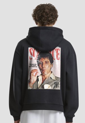Fekete kapucnis pulóver nagy grafikával a hátulján. A képen egy férfi látható, aki cigarettát tart, alatta szöveg található. Puha, texturált anyag.