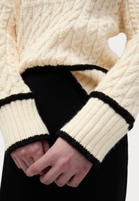 Maglione color crema a lavorazione a trecce con maniche a costine, con rifiniture nere. In primo piano, mani che tengono il maglione in vita.