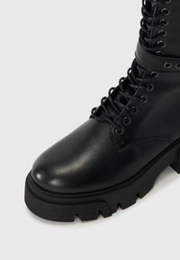 Juodas odinis kulkšninis botas su lygiu paviršiumi, apvaliu nosiu, tvirtais raišteliais ir masyviu platformos padu su dantyta kraštovaizdžio apdaila.