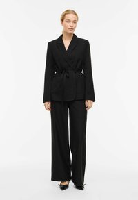 Blazer nero su misura con maniche lunghe, cintura da annodare in vita e due tasche frontali, abbinato a pantaloni neri a gamba larga e scarpe nere con punta affilata.