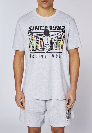Mann trägt hellgraues Sportoutfit mit Grafik einer Person mit ausgestreckten Armen und Text „SINCE 1982 Active Wear“ auf dem Shirt.