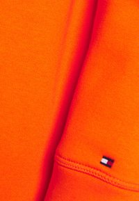 Heldere oranje trui met een gladde textuur, voorzien van geribbelde manchetten en een klein logoplaatje in rood, wit en blauw op de mouw.
