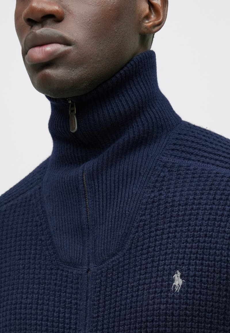 Suéter azul marino de punto con cremallera y cuello alto, con tejido texturizado y un pequeño logo blanco en el pecho. Detalle de cremallera en el cuello.