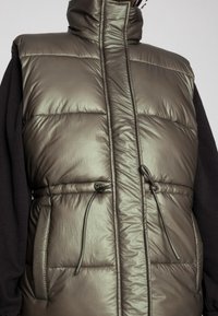 Padded vest w metalicznej oliwkowej zieleni, z wysokim kołnierzem, regulowanym ściągaczem w talii i kieszeniami bocznymi. Gładka, teksturowana powierzchnia.