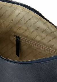 Interior texturizado de color azul oscuro de una bolsa que presenta un bolsillo con cremallera y un forro color beige con el logotipo repetido "HARBOUR 12ND". Detalle de cremallera de metal visible.