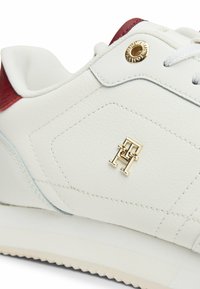 Witte leren sneaker met een rode accent bij de hak, met een gouden logo detail en een gestructureerde stiknaad. Rubberen zool met profiel voor grip.