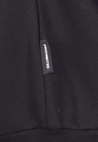 Tessuto nero con una texture liscia, caratterizzato da un'etichetta con la scritta "PROIBITO" in bianco cucita nella cucitura laterale.
