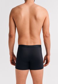 Schwarze Boxershorts mit einem weichen elastischen Bund, tailliertem Design und glatter Textur, von hinten auf einem neutralen Hintergrund betrachtet.