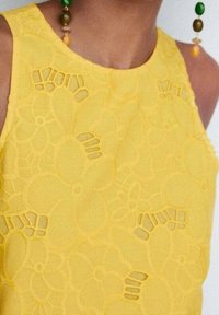 Żółty top bez rękawów z kwiatowym wzorem koronkowym, zawierający wycięte motywy kwiatów i gładką teksturę. Uzupełniony kolorowymi kolczykami z koralikami.