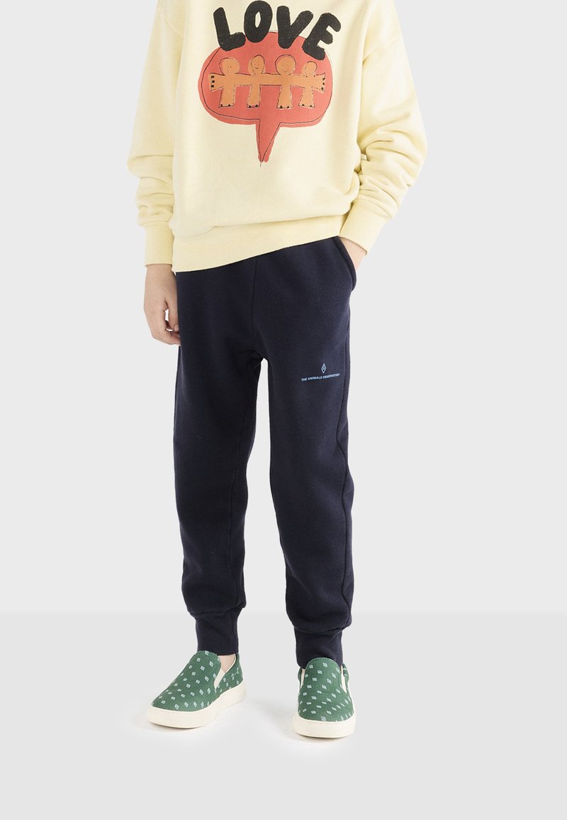 Gelbes Sweatshirt mit "LOVE" Grafik, marineblaue Sweatpants mit Seitentaschen und grüne Slipper mit weißen, gepunkteten Akzenten.