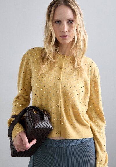 Κίτρινο κατεργασμένο με πασμένες πέρλες, σ�τρογγυλή λαιμόκοψη και κλείσιμο με κουμπί, συνδυασμένο με μια σκούρα υφαντή τσάντα και τυπωμένη μπλε φούστα.