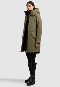 Olivgrüne Daunenjacke mit hohem Kragen, Reißverschluss, zwei seitlichen Taschen und schwarzen Akzenten; kombiniert mit schwarzen, engen Hosen und kniehohen Stiefeln.