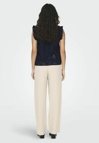 Top ricamato blu navy con maniche a volant, abbinato a pantaloni beige chiaro a gamba larga e scarpe nere, visto da dietro.