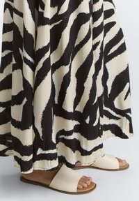 Schwarzes und cremefarbenes, gepunktetes Maxikleid mit Zebra-Streifen; verfügt über ein fließendes Design und wird mit hellen Slide-Sandalen auf einer flachen Oberfläche kombiniert.