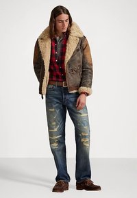 Brązowa kurtka ze skóry z kożuchem i jasnym kołnierzem, czerwona i czarna koszula w kratę oraz przetarte niebieskie dżinsy z postrzępionymi detalami.