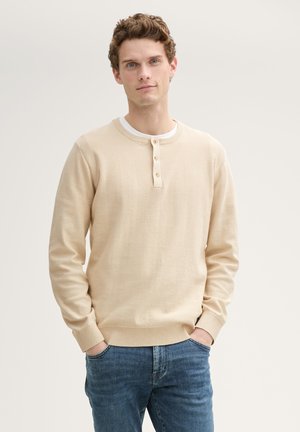 Pull beige tricoté à manches longues avec un col rond et une patte de boutonnage à trois boutons, associé à un jean bleu. Texture lisse et couleur neutre.