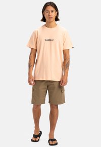 Camiseta de algodón de color rosa claro con un logo de Quiksilver, combinada con pantalones cortos cargo color caqui y chanclas negras. Diseño simple con mangas cortas.