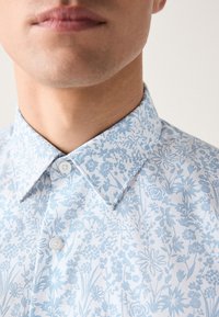 Világoskék, virágmintás ing hegyes gallérral, gombos lezárással és apró, részletes virágmintákat ábrázoló texturált anyagból.