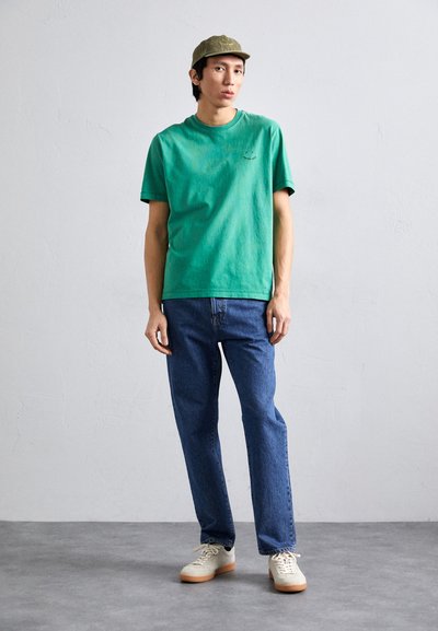 Πράσινο βαμβακερό μπλουζάκι με κοντά μανίκια, μπλε τζιν παντελόνι και μπεζ αθλητικά παπούτσια. Το μοντέλο φοράει ένα ανοιχτό πράσινο καπέλο. Απλό φόντο.