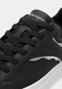 Zapatilla de cuero negra con superficie texturizada, que presenta cordones negros, acentos plateados y una suela de goma blanca. El diseño notable incluye un patrón ondulado.