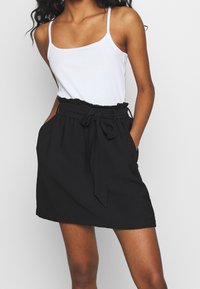Jupe noire avec une taille nouée, dotée de poches latérales et d'une coupe décontractée. Associée à un débardeur blanc, présentant un design simple.