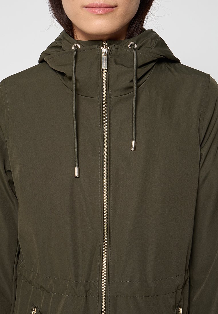 Kobieta w oliwkowej kurtce z kapturem, z zamkiem błyskawicznym z przodu i sznurkiem do regulacji, ujęta od ramion do ust na jednolitym tle.