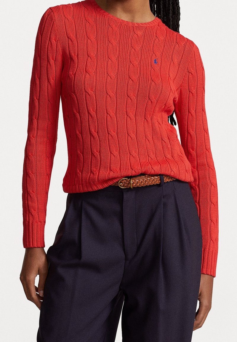 Femme portant un pull rouge à torsades rentré dans un pantalon plissé bleu marine avec une ceinture tressée marron, debout avec une main détendue.
