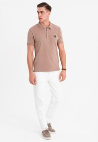 Svetle hnědý polo triko s límcem na zip a malým logem, v kombinaci s bílými kalhotami a hnědými semišovými botami. Minimalistický design.