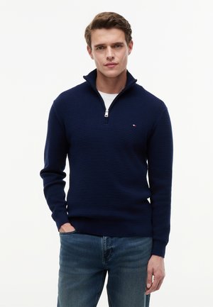 Man with short brown hair wearing a navy blue textured quarter-zip sweater over a white shirt and blue jeans, standing with one hand in pocket.