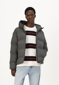 Grå polstret jakke med hætte, der har en glat tekstur. Båret over en stribet striktrøje i hvid, rød og marineblå. Lysgule jeans.