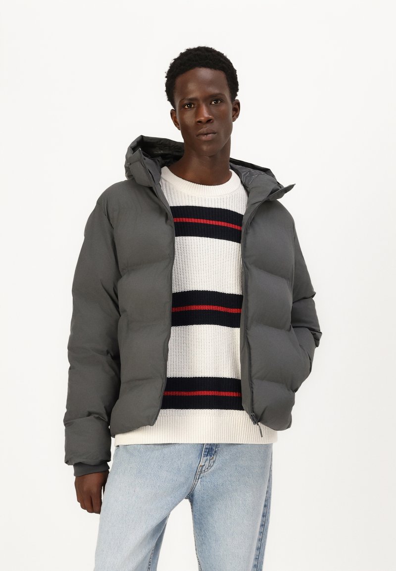 Grå polstret jakke med hætte, der har en glat tekstur. Båret over en stribet striktrøje i hvid, rød og marineblå. Lysgule jeans.