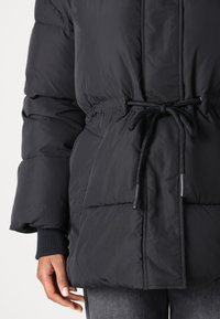 Black puffer jacket with a drawstring waist, features quilted texture and ribbed cuffs. Large front pockets and a high collar.