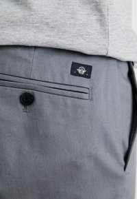 Pantaloni in cotone grigi con chiusura a bottone, tasche laterali e un'etichetta in tessuto scuro con logo. Texture liscia con dettagli di cucitura visibili.