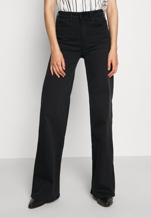Kvinde iført højtaljede, løstsiddende sorte jeans kombineret med en sort-hvid stribet top og sorte spidse sko.