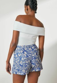 Top alb cu decolteu off-the-shoulder, cu un model texturat, asortat cu șorturi albastre cu imprimeu floral și un design relaxat.