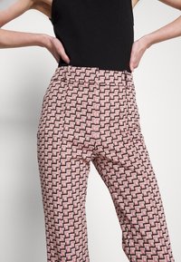 Femme portant un pantalon taille haute rose à motif géométrique noir et blanc et un haut noir sans manches, les mains sur les hanches.