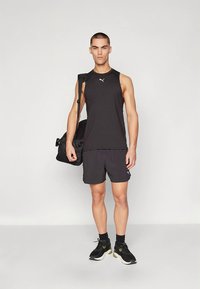 Černé atletické tílko a šortky, obojí s jemným logem. Modelka nese černou sportovní tašku a má na nohou černé běžecké boty se zelenými akcenty.