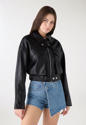 Giovane donna che indossa una giacca di pelle nera testurizzata e pantaloncini di jeans blu asimmetrici, in piedi davanti a uno sfondo chiaro e semplice.