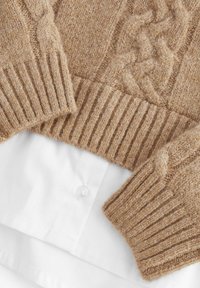 Beige kötött pulóver, texturált fonott mintával és bordázott mandzsettákkal, fehér galléros ing fölé rétegezve, gombbal.