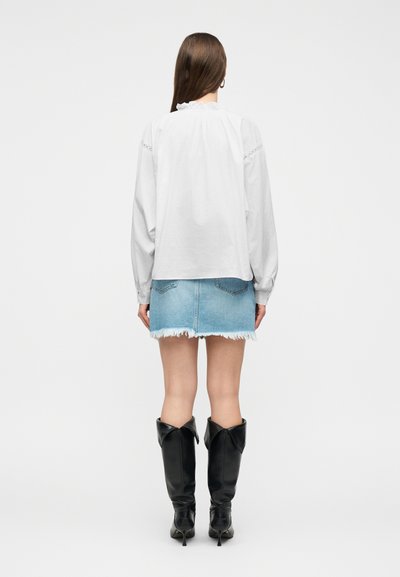 Γυναίκα με μακριά καστανά μαλλιά φοράει λευκό πουκάμισο, φούστα από ανοικτό μπλε ξεφτισμένο τζιν και μαύρα μπότες με τακούνι που φτάνουν μέχρι το γόνατο, γυρισμένη με την πλάτη.