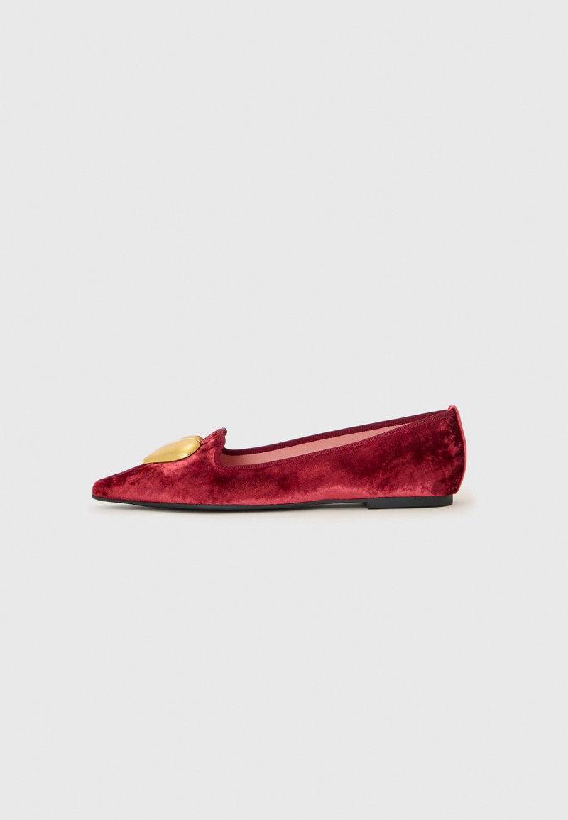 Pantofii plat roșii din catifea, cu vârf ascuțit, prezintă o embellisment aurie pe partea din față și o talpă neagră. Textură netedă și design elegant.