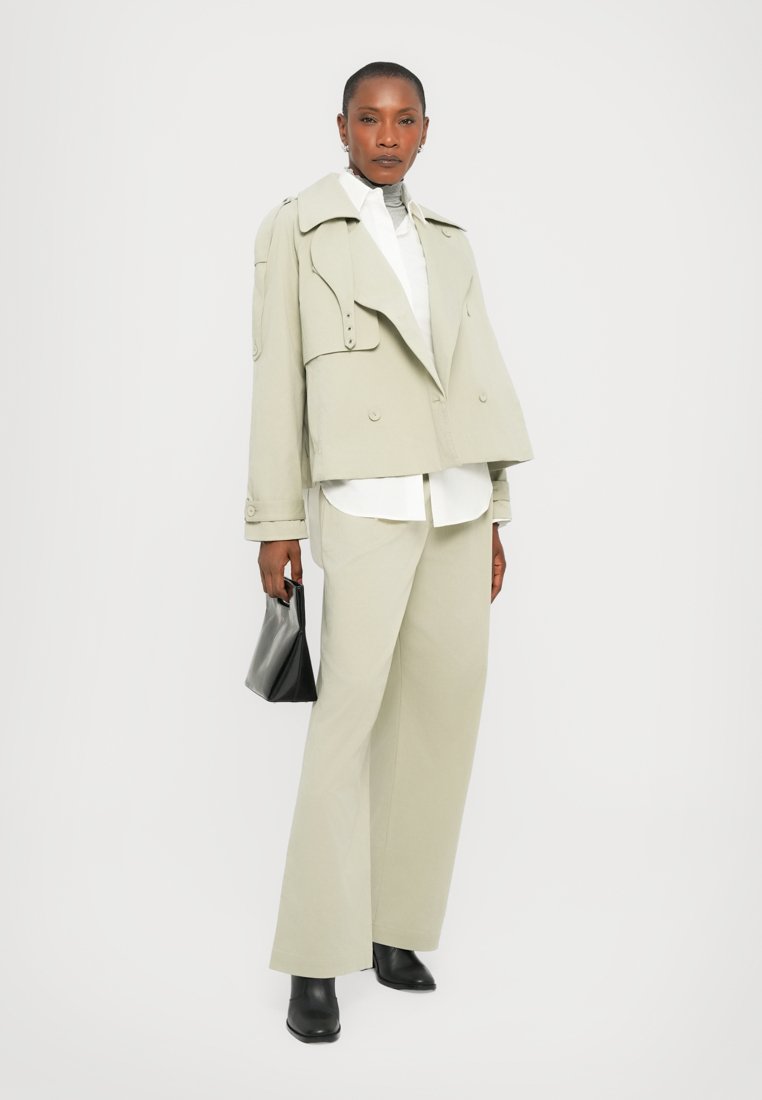 Veste courte gris clair avec large revers et accents de boutons, associée à une chemise blanche et un pantalon assorti. Un sac à main noir et des bottes complètent le look.