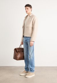 Chaqueta de forro polar beige con dos bolsillos frontales, camiseta beige clara, vaqueros azules y zapatillas color beige; llevando un bolso de piel marrón.